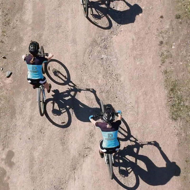 Tre personer cyklar på en grusväg omgiven av grönska under en klarblå himmel.